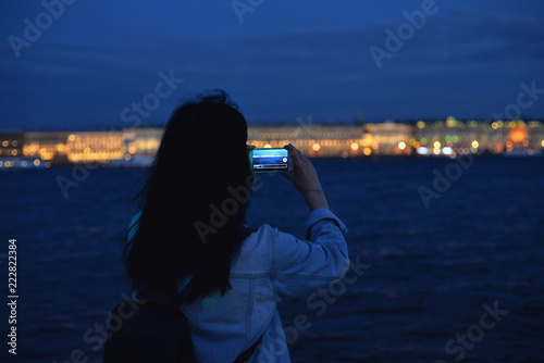 a tourist in a denim jacket holds a smartphone and takes pictures of the night St. Petersburg. A beautiful view from the side of the Petropavlovsk fortress on the Neva and the night lights