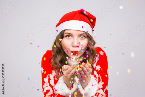 Holiday, Christmas and people concept - Beautiful and young woman in santa claus costume over white background blowing on confetti
