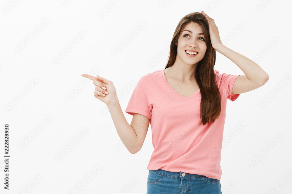 Indoor shot of good-looking dreamy girlfriend in casual outfit, holding palm on head and gazing up with beautiful and tender smile while pointing left, remembering something over grey wall Indoor shot of good-looking dreamy girlfriend in casual outfit, holding palm on head and gazing up with beautiful and tender smile while pointing left, remembering something over grey wall