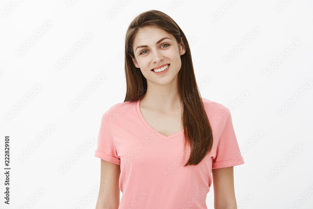 Friendly good-looking female coworker in pink t-shirt smiling broadly and tilting head, listening or talking with customer, being polite and in good mood while standing against gray background Friendly good-looking female coworker in pink t-shirt smiling broadly and tilting head, listening or talking with customer, being polite and in good mood while standing against gray background