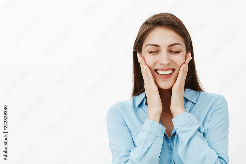 Portrait of pleased charming european brunette in blue blouse, closing eyes and smiling broadly, touching cheeks gently, blushing after perfect date with romantic boyfriend, standing over gray wall Portrait of pleased charming european brunette in blue blouse, closing eyes and smiling broadly, touching cheeks gently, blushing after perfect date with romantic boyfriend, standing over gray wall