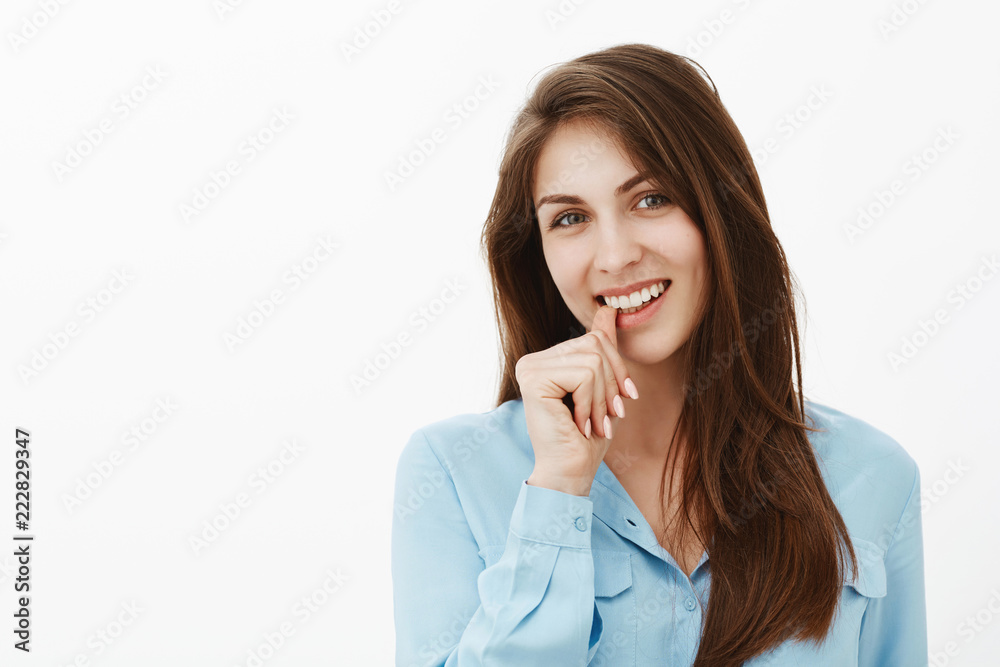 Flirty charming female employee in blue blouse, smiling broadly and biting thumb while gazing at camera with intrigued and interested expression, having great idea or intention over gray wall Flirty charming female employee in blue blouse, smiling broadly and biting thumb while gazing at camera with intrigued and interested expression, having great idea or intention over gray wall