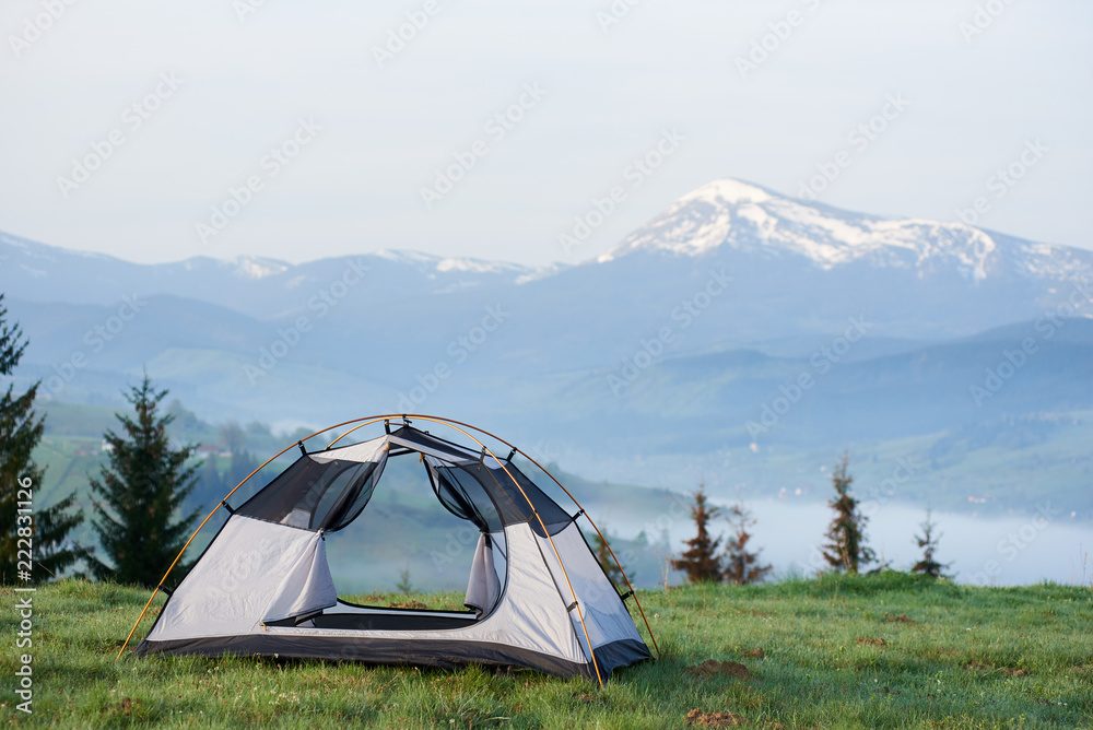 Open empty small tourist tent on grassy hill on background of pine trees and distant mountain range with snowy peaks lit by morning sun. Tourism, camping, recreation and beauty of nature concept.
