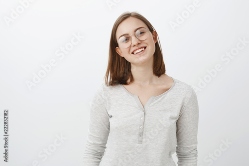 Waist-up shot of smart creative charming young woman with short cute haircut tilting head joyfully smiling broadly wearing glasses and casual white blouse posing happily over grey background