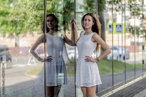 Fototapeta Naklejka Na Ścianę i Meble -  Beautiful young girl in a white summer dress near a showcase with reflection
