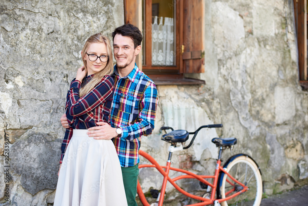 Portrait of young attractive tourist couple, bearded man and blond woman standing together, smiling and looking to the camera at modern tandem bicycle on empty pavement at cracked wall of old building
