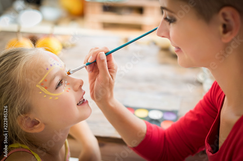 Young mother painting daughters face for Halloween party. Halloween or carnival family lifestyle background. Face painting and dressing up.