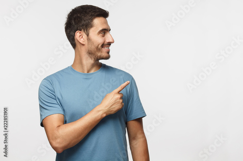 Casual dressed young man in blue t-shirt looking right, pointing with his finger, isolated on gray background with copy space for your text