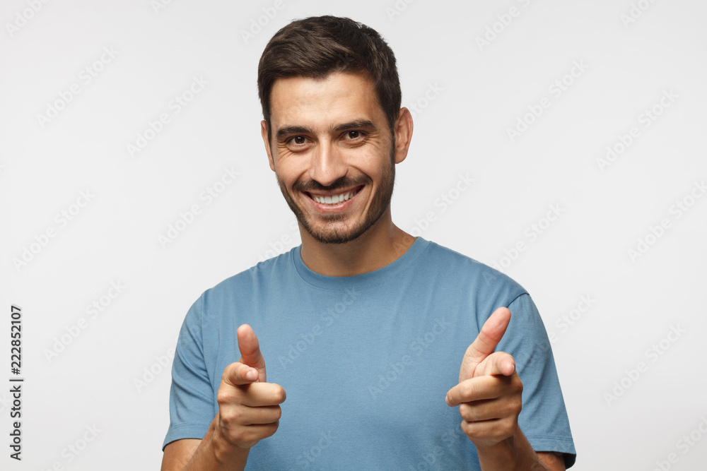 Young man isolated on gray background wearing blue t-shirt, smiling happily with open mouth and pointing to camera with both hands Young man isolated on gray background wearing blue t-shirt, smiling happily with open mouth and pointing to camera with both hands
