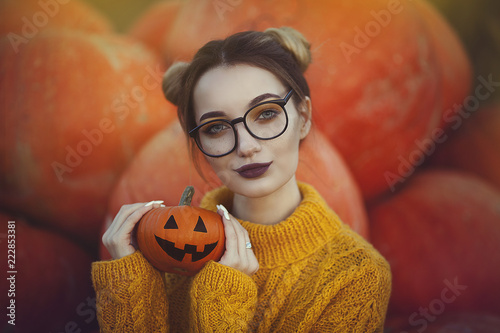 A cozy autumn photo of a girl with pumpkins in a yellow sweater and glasses. Stylish young woman in autumn decorations, in hands a Halloween pumpkin.