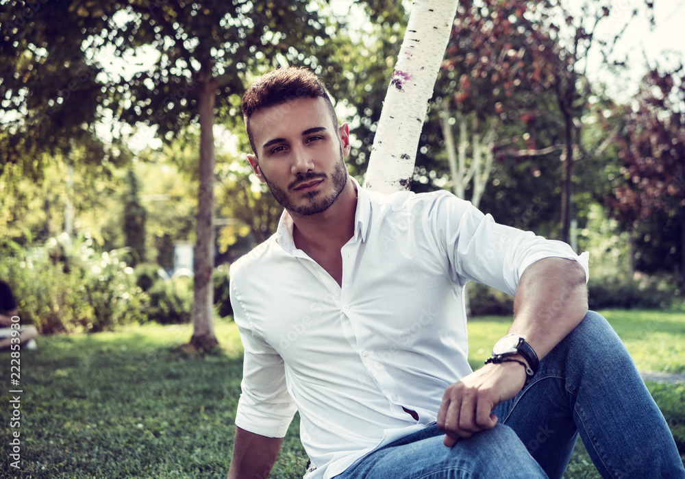Attractive handsome young man in city park, sitting, in a summer day, wearing jeans and white shirt Attractive handsome young man in city park, sitting, in a summer day, wearing jeans and white shirt