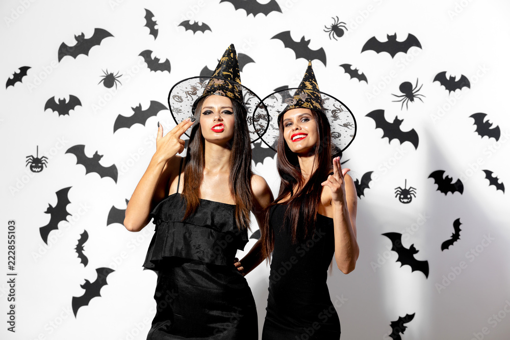 Two brunette women in black dresses and witches hats with red lipstick have fun on a white background with bats and spiders Two brunette women in black dresses and witches hats with red lipstick have fun on a white background with bats and spiders