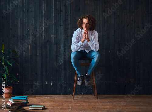 Handsome curly student holds hands on chin while sitting on a chair in a room with the minimalist interior.