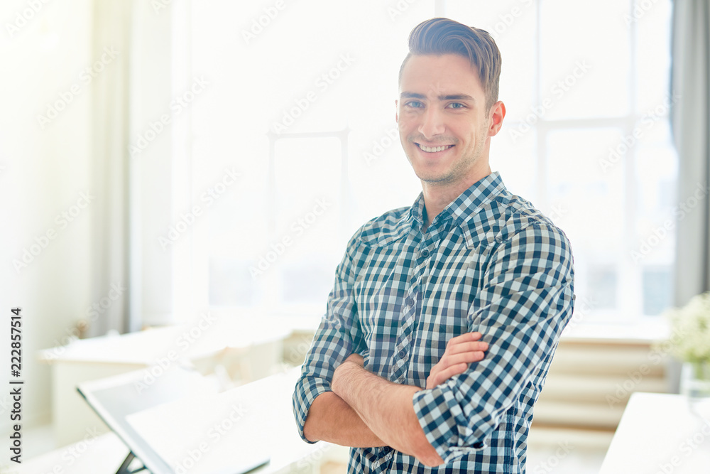 Cross-armed guy in checkered shirt standing in white studio full of sunlight