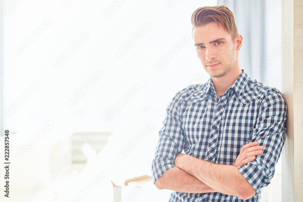 Serious guy with his arms crossed on chest looking at you while leaning against wall of modern design studio Serious guy with his arms crossed on chest looking at you while leaning against wall of modern design studio