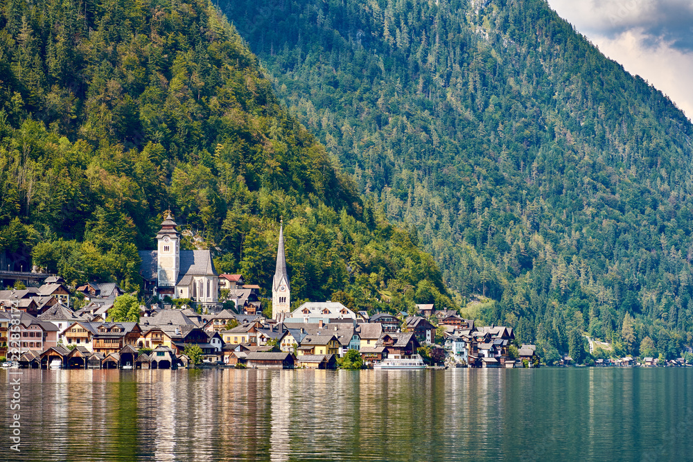 View of the lake and the city of Hallstatt with church, mountains and forests, under the sky with clouds. Reflection on water.