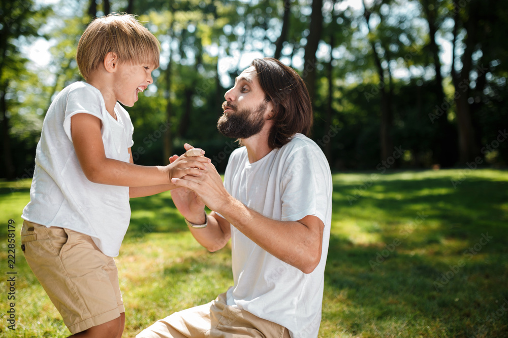 Dark-haired dad and his son dressed in white t-shirts fool around and having fun on the green lawn in the park on a sunny day Dark-haired dad and his son dressed in white t-shirts fool around and having fun on the green lawn in the park on a sunny day