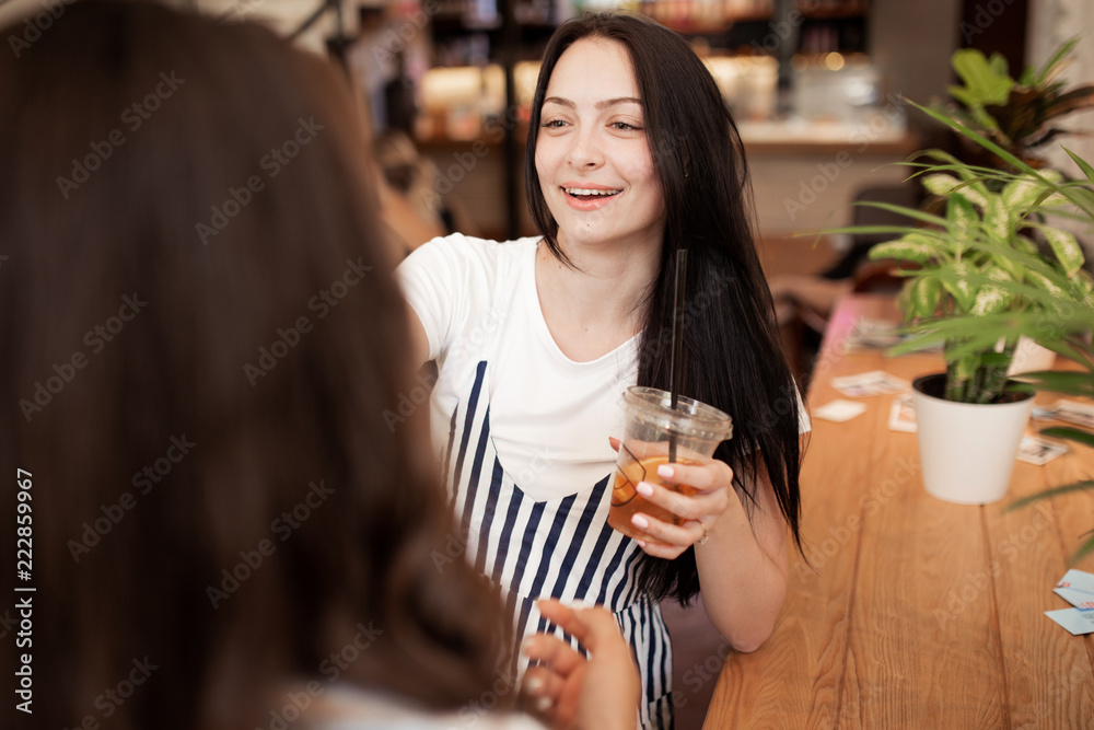Two beautiful youthful smiling girls with dark hair,dressed in casual outfit,stand opposite each other and chat in a cozy coffee shop.