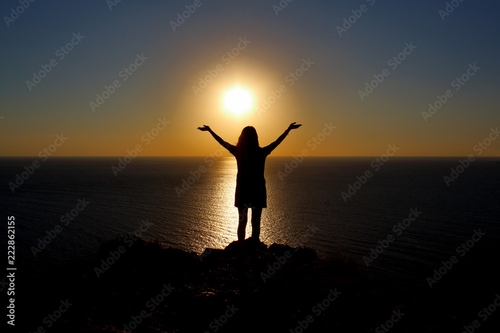 young girl is facing the sun on the mountain and looking at the sea, hands raised up and to the side