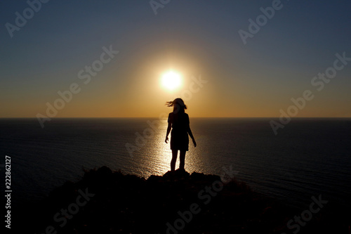 young girl is facing the sun on the mountain and looking at the sea, hands raised up and to the side