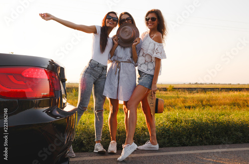 Three beautiful young girls stand in the field next to the car and talk on a warm sunny day.