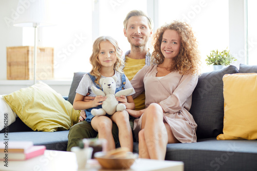 Cute girl with teddybear sitting on her dad knees with mom near by while the family relaxing at home