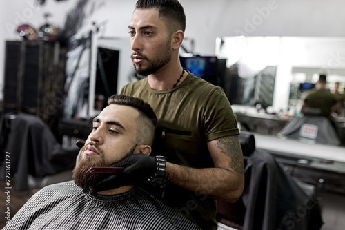 Handsome barber holds his hands on the beard of young brutal man and looks at the mirror at a barbershop