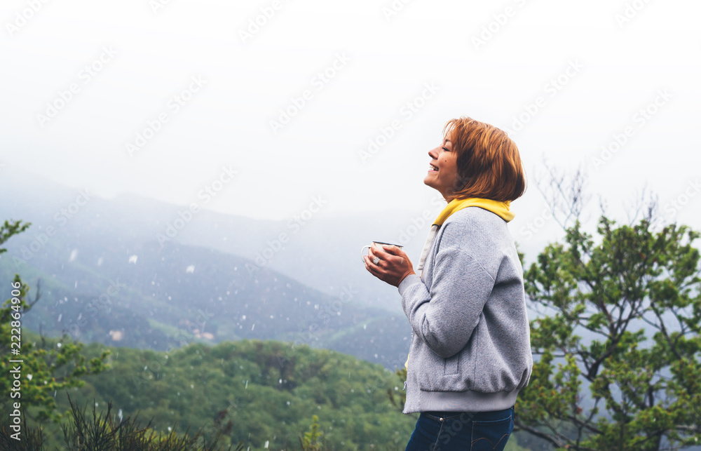 tourist traveler standing on green top on mountain, girl smiling and holding cup with drink against a background of falling snow on hills, happy girl enjoying nature panoramic landscape, relax concept tourist traveler standing on green top on mountain, girl smiling and holding cup with drink against a background of falling snow on hills, happy girl enjoying nature panoramic landscape, relax concept