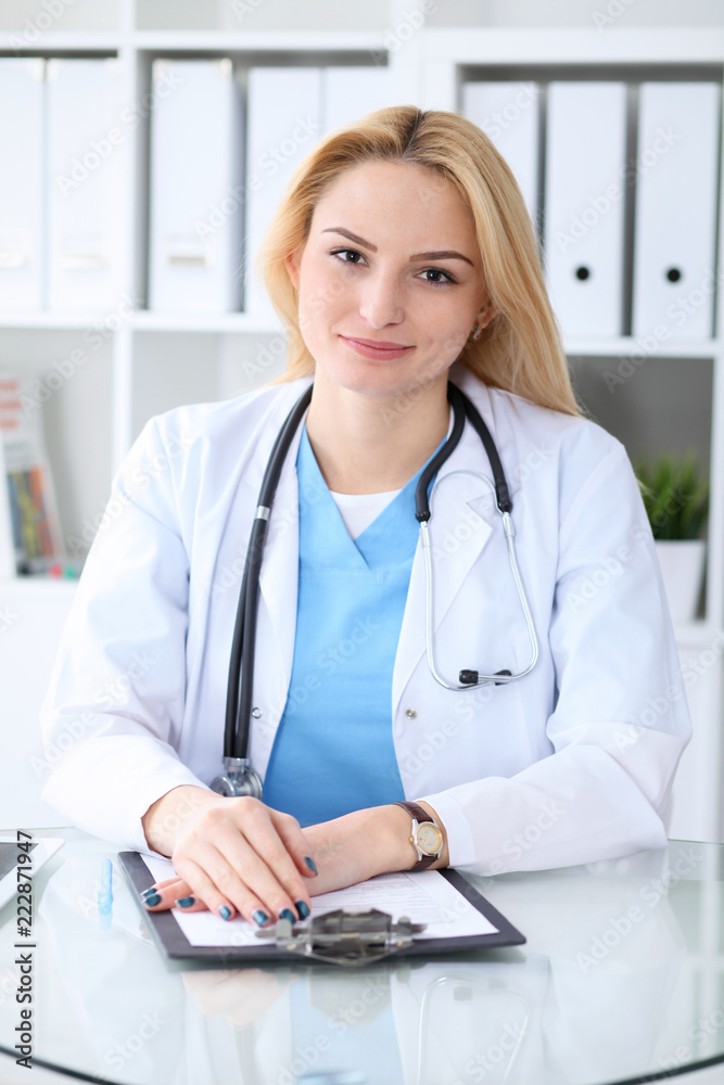 Doctor woman at work. Portrait of cheerful smiling blonde physician sitting at the desk. Medicine and healthcare concept