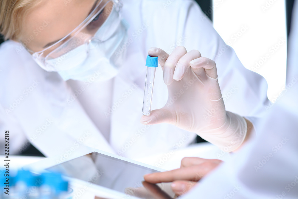 Closeup of scientific research team with clear solution in laboratory. Blonde female chemist holds test tube of glass while her colleague checks results with tablet pc. Blood test, medicine or