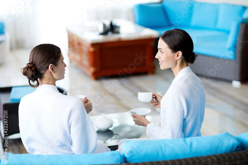 Two girls in white bathrobes sitting on sofa and talking by cup of tea or coffee in spa center
