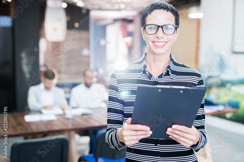 Professional woman in office with eyeglasses and confident expression as other workers hold a meeting in background © Seventyfour