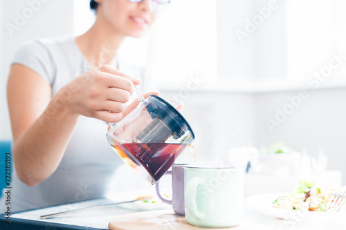 Closeup of unrecognizable young woman pouring black tea from glass kettle while enjoying lunch in sunlit cafe, copy space