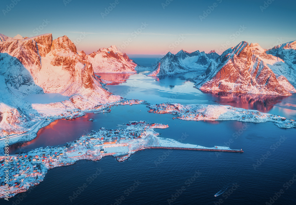 Aerial view of Reine at sunrise in winter. Top view of Lofoten islands, Norway. Panoramic landscape with blue sea, snowy mountains, high rocks, village with buildings, rorbu, sky reflected in water