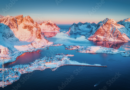 Fototapeta Naklejka Na Ścianę i Meble -  Aerial view of Reine at sunrise in winter. Top view of Lofoten islands, Norway. Panoramic landscape with blue sea, snowy mountains, high rocks, village with buildings, rorbu, sky reflected in water
