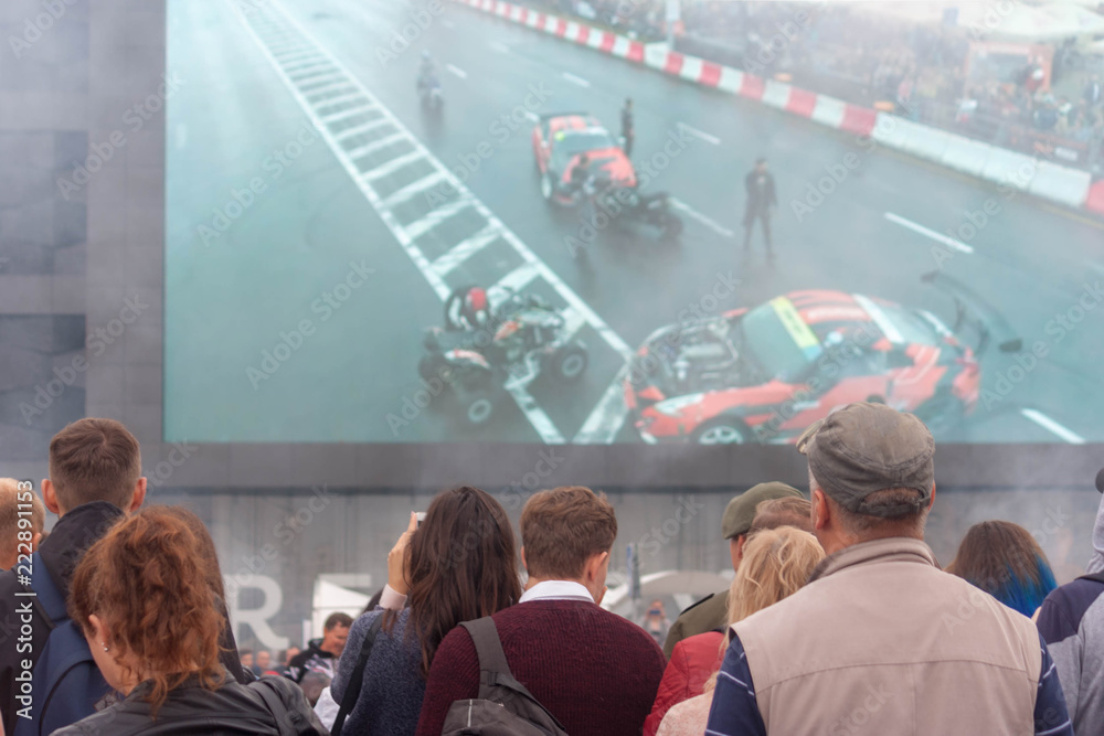 A view of a number of spectators who watch the motor show on the background of a large screen that broadcasts the spectacle in real time.