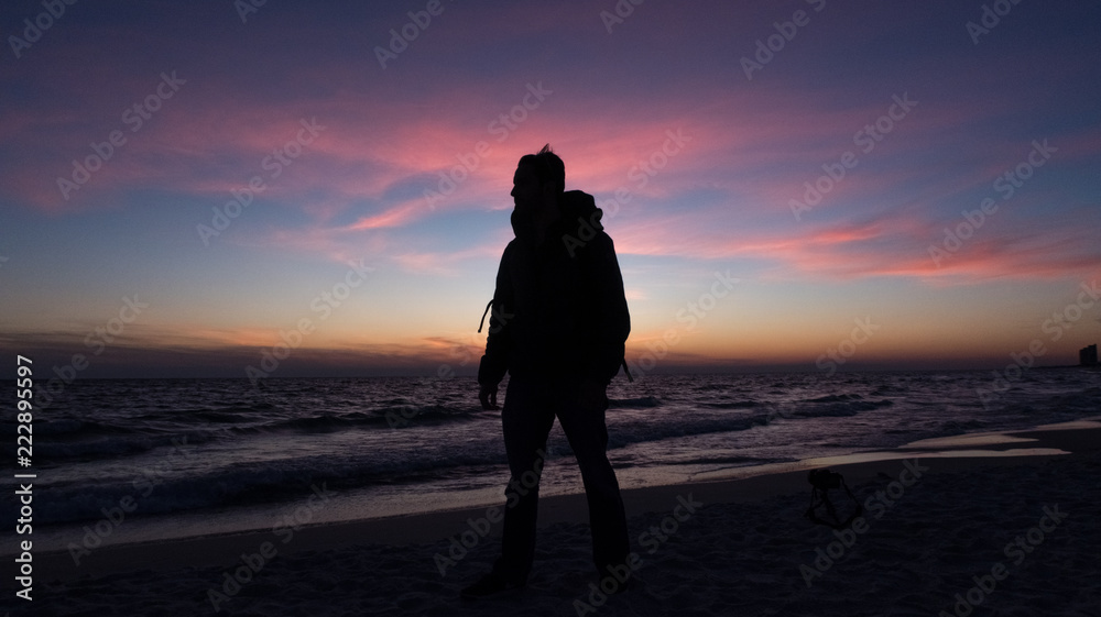 Guy stands in front of dramatic sunset over the ocean on a beach covered with waves.