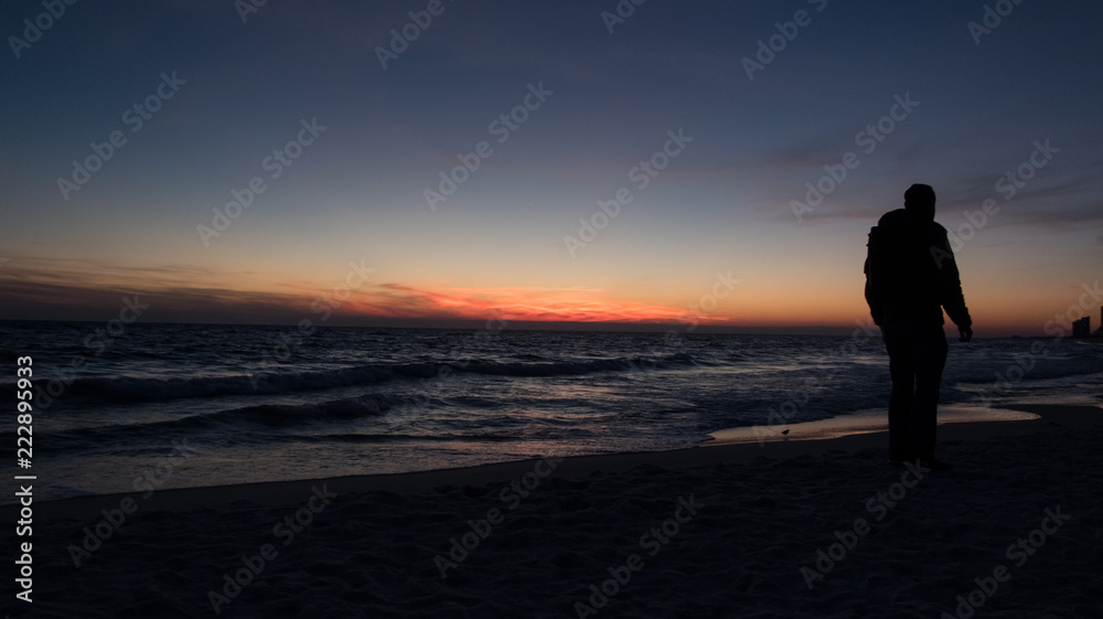 Guy stands in front of dramatic sunset over the ocean on a beach covered with waves.