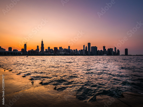 Chicago skyline picture during a beautiful sunset with purple and orange sky above and building silhouettes with rippling waves and water of Lake Michigan and concrete shoreline in the foreground.
