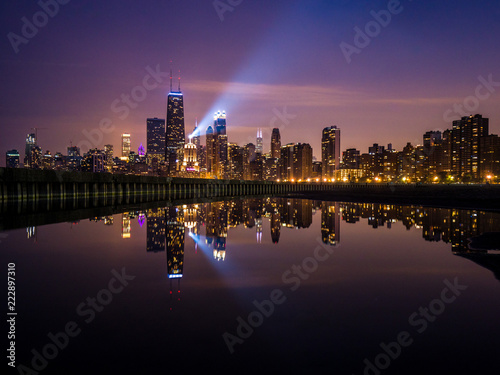 Beautiful long exposure Chicago night skyline photo with building lights at sunset with pink purple and blue clouds in the sky reflecting in a calm pool of water and spotlight shining up into the sky.