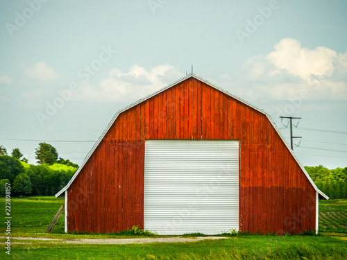 Beautiful photograph of a freshly painted red vintage weathered wood barn with pitched roof in a field with blue sky and white fluffy clouds above.