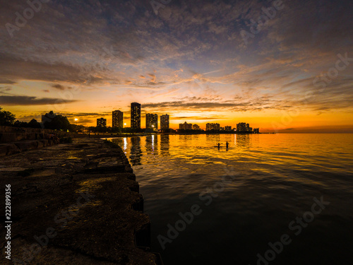 Gorgeous sunset photograph looking across Lake Michigan at Foster Beach to the highrise buildings and colorful sky on the horizon as kids play and swim in the water near the shoreline.