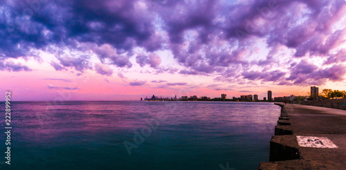 Beautiful panorama photograph of the Chicago skyline at sunset with fluffy purple and pink clouds in the sky above and blue water below along the concrete shoreline at Montrose Beach.