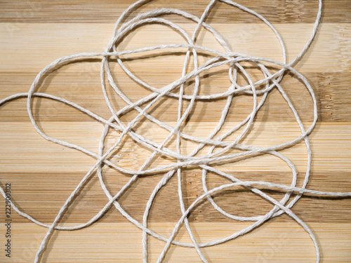 Close-up of white braided roll of thin white string or yarn unraveled and tangled laying randomly on top of a bamboo wood cutting board background.