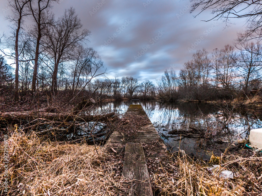 View looking down a wood plank to a weathered pier sticking out into the calm water of a pond at sunset with pink and blue fluffy clouds above and trees reflecting in the water. View looking down a wood plank to a weathered pier sticking out into the calm water of a pond at sunset with pink and blue fluffy clouds above and trees reflecting in the water.