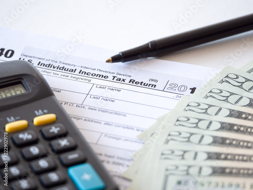 Close-up photograph of a hard copy of an individual income tax return with black calculator and 20 dollar bills piled up representative of United States federal tax cuts and savings this year.