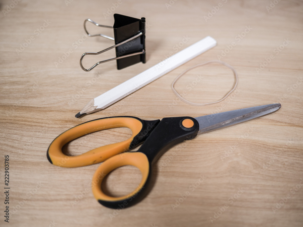 Close up photograph of office supplies including a scissors, rubber band, a white carpenter pencil, and a black metal binder clip on top of a light colored wood grain desk. Close up photograph of office supplies including a scissors, rubber band, a white carpenter pencil, and a black metal binder clip on top of a light colored wood grain desk.