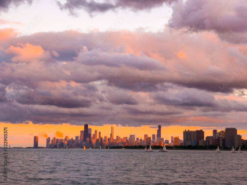 Gorgeous landscape panoramic photograph of the beautiful Chicago skyline with buildings at horizon and Lake Michigan in foreground and pink, orange, and purple fluffy clouds in sky above.