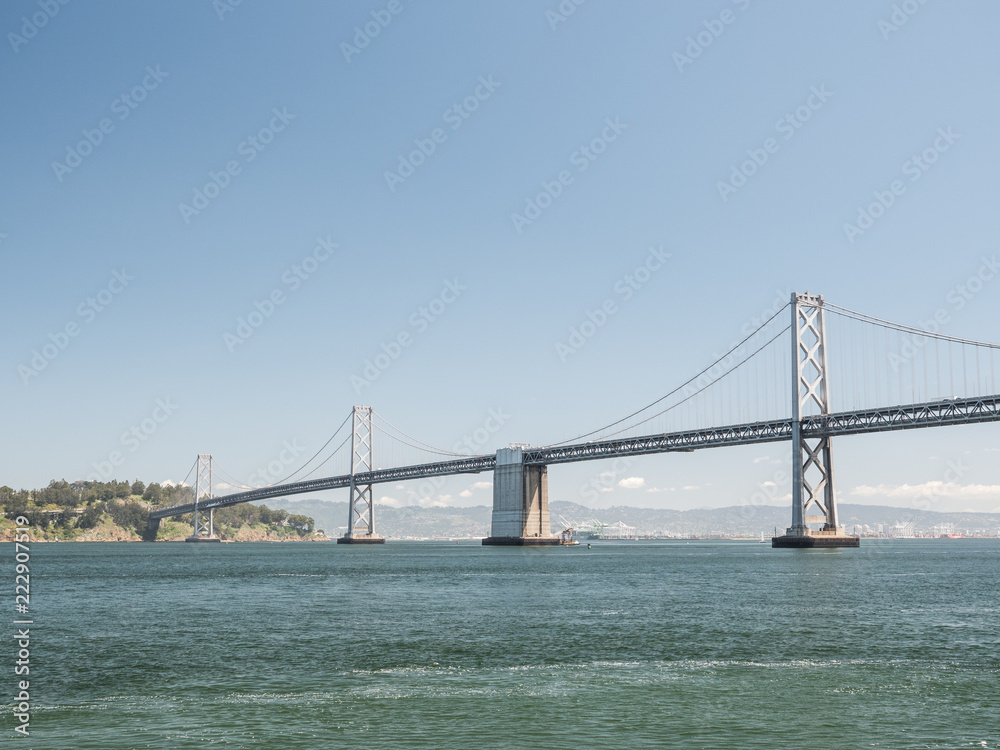 A beautiful view of the expensive and famous San Francisco Oakland Bay Bridge with suspension cables and towers protruding from the San Francisco Bay with water below and clear blue sunny sky above.