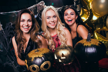 Which pumpkin is more scary? Beautiful young women in evening gown looking at camera and holding pumpkins with smile while celebrating Halloween in nightclub
